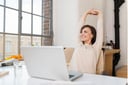 A horizontal image. A woman in a light-colored sweater sits at a table, leaning back and stretching her arms above her head. She smiles broadly, showing her teeth, and looks out of the window to her right. The view outside the window is partially cropped out of the frame, showing only a pale sky and part of the wall of a neighboring building. An open laptop sits on the table in front of her.