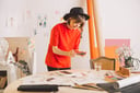 Horizontal image. A woman in an art studio wearing a red shirt and a black wide-brimmed hat. She is leaning over a table, taking a photo of the paintings on the table.