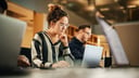 Horizontal image: A woman is looking at a laptop screen on a table. The laptop is facing the viewer, so the screen is not visible. The woman has curly hair, a striped shirt, and glasses. To her left is a man wearing glasses, but he is blurred and difficult to see clearly.